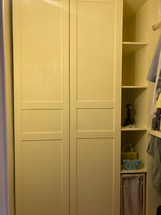 Gul garderob med dubbeldörrar intill en vägg. Garderoben ser sned ut jämfört med väggen. På sidan finns öppna hyllor med diverse föremål.