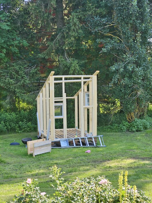 Byggstomme till mindre byggnad påbörjad på gräsmatta med skog i bakgrunden. Verktyg och material ligger på marken runtom.