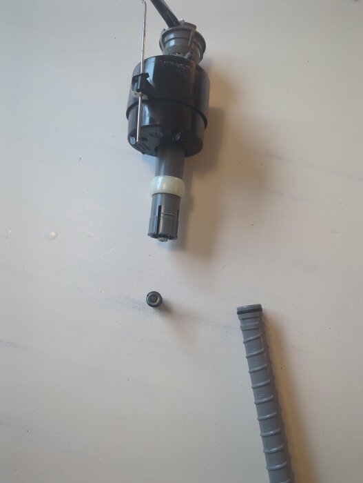 En isärplockad flottör från en äldre Gustavsberg-toalett, med en svart plastdel, ett grått rör och en liten gummidel placerade på ett vitt underlag.