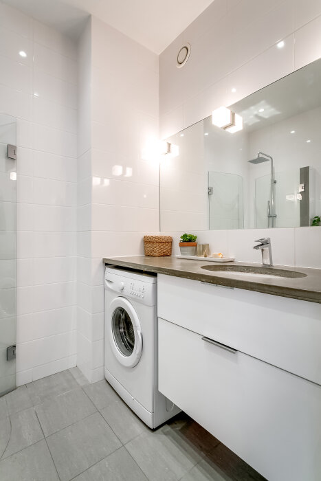 Modernt badrum med tvättmaskin under en grå bänkskiva, handfat, stor spegel och dusch. Mysiga detaljer som krukväxter och korgar. Välbelyst och stilrent.