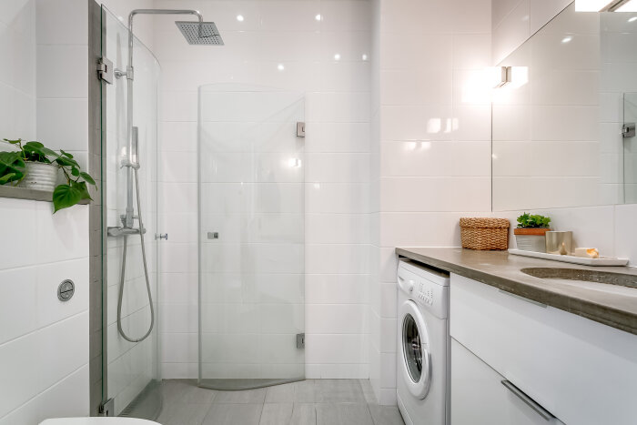 Modernt badrum med dusch, tvättmaskin, väggskåp och bänk. Gröna växter och korgar som dekorationer. Vitt kakel på väggarna och ljusgrått klinker på golvet.