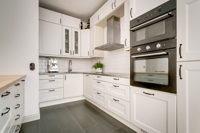 Nyrenoverat vitt kök med svarta detaljer, inbyggd dubbelugn, fläkt, diskbänk, och kaffemaskin. Växt på diskbänken och vita kakelväggar.