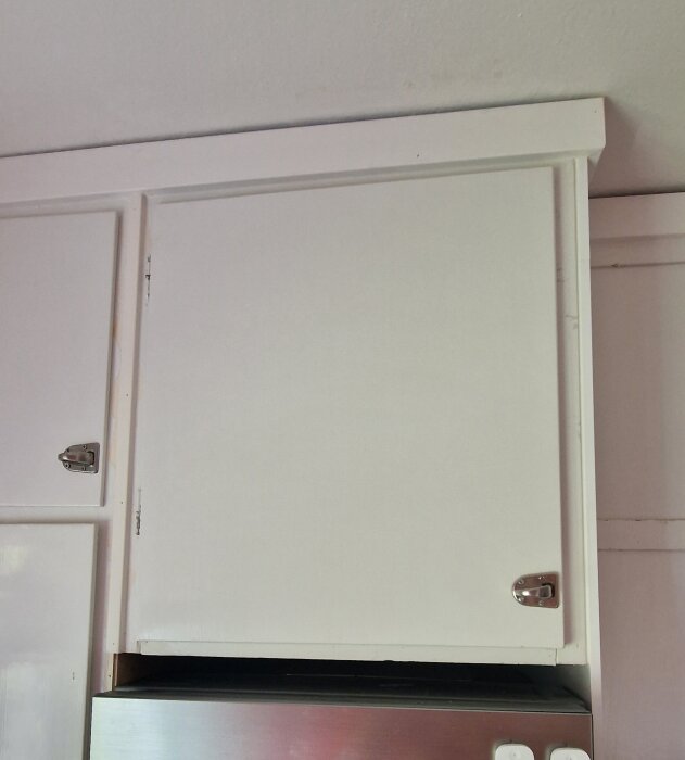 Vitt överskåp i köket, del av omlokaliserat kylskåpsuttag syns till höger ovanför den rostfria kylen. Skåpets botten och dörr har höjts.