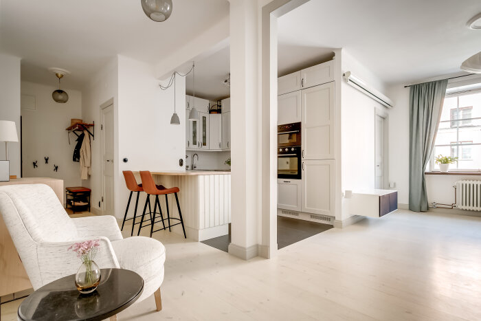 Ljus och modern inredning i en lägenhet med öppet kök, vita väggar och trägolv. En fåtölj och bord med en blomvas står i förgrunden.
