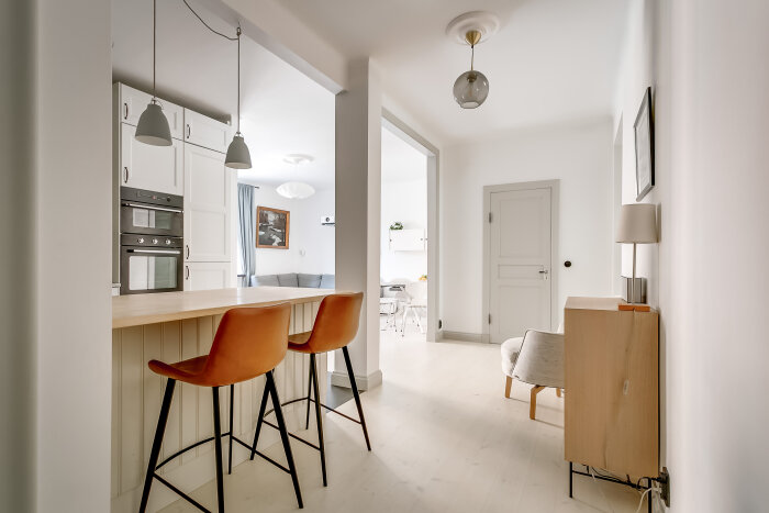 Modern köksö med två orange stolar, vita hängande lampor, vita skåp och dubbla varmluftsugnar, öppet mot vardagsrum och matplats inredning.