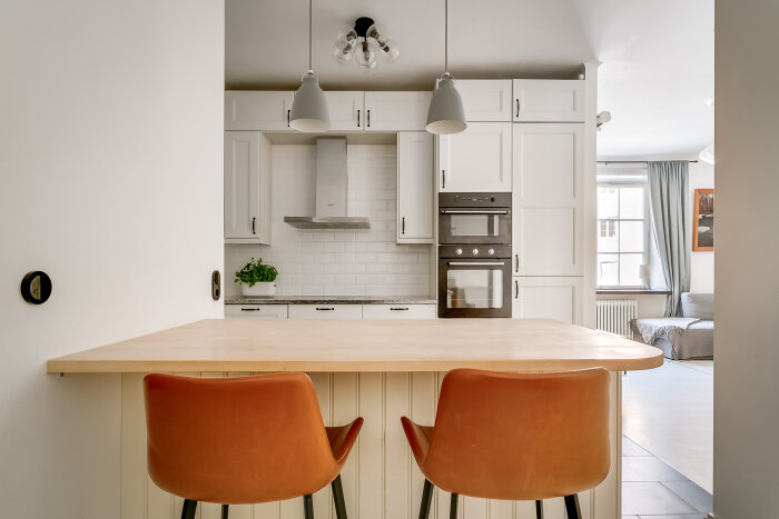 Modernt kök med vita skåp, rostfri spisfläkt, inbyggd ugn, två orangea stolar vid köksön, och ett fönster med gardiner i bakgrunden.