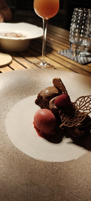 Närbild på en dessert med choklad, hallon, och ett kulglass samt en orange drink och ett glas vatten på ett bord.