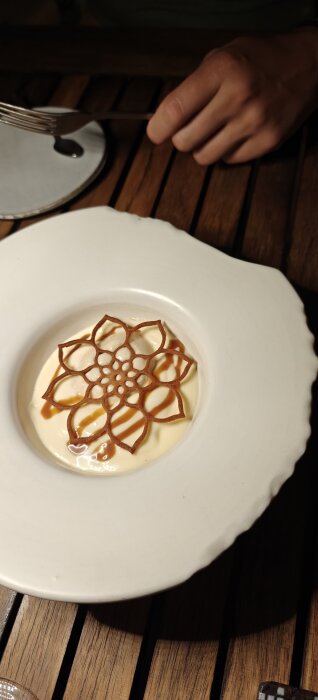 En dessert på ett vitt porslinsfat bestående av glass och sås, toppad med ett dekorativt, blommönstrat krisp. En hand håller en gaffel bredvid tallriken.