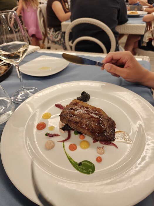En tallrik med en stekt köttbit och dekorativa såsfläckar, ett glas vin och en hand som håller en kniv. Bakgrund med andra restauranggäster.