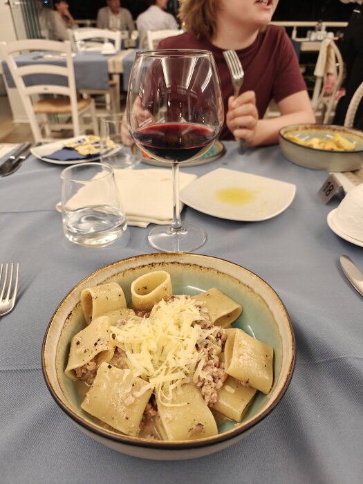 En tallrik med pasta toppad med ost bredvid ett glas rödvin på ett restaurangbord. En person i röd tröja håller en gaffel i bakgrunden.
