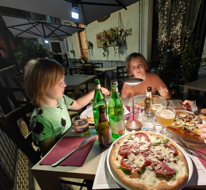 Två barn som sitter vid ett bord på en restaurang i Sicilien. På bordet finns pizza, ölflaskor och glas med dryck.
