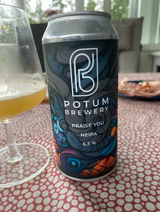 En burk Potum Brewery Praise You NEIPA och ett glas öl står på ett bord med röd- och vitmönstrad duk, med plockmat i bakgrunden.