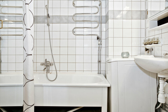 Renoverat badrum med vita kakelplattor, badkar med duschdraperi, handfat, tvättmaskin och handdukstork.