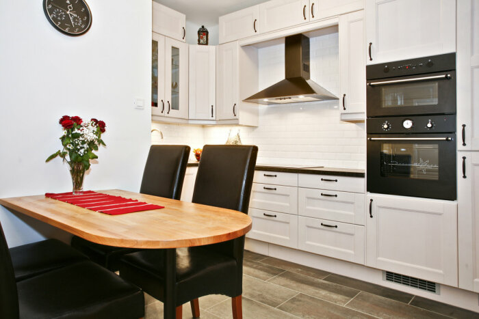 Vit och modern köksrenovering med vita skåpluckor, svart köksfläkt och ugn. Matbord med röda rosor och klocka på väggen.