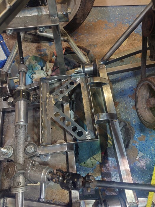 Närbild på en halvmonterad stålram och en mekanisk del i ett garage med verktyg och trasor i bakgrunden.