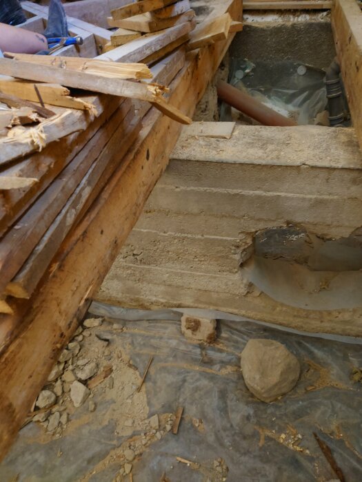 Rivningsarbete av badrum visar synliga bjälkar, använd verktyg och stokar. Betongvägg med ett rödorange rör och uppbruten yta. Sten och träbitar på golvet.