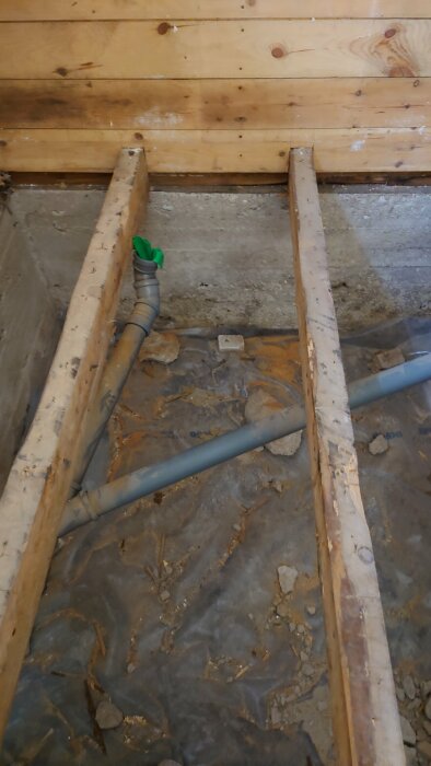 Badrum under renovering där golvet är rivet, synliga bjälkar, en avloppsledning och en betongvägg i bakgrunden.
