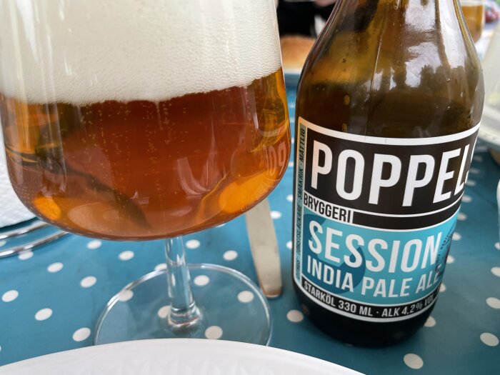 En flaska Poppels Bryggeri Session India Pale Ale bredvid ett glas öl med skum på en prickig duk.