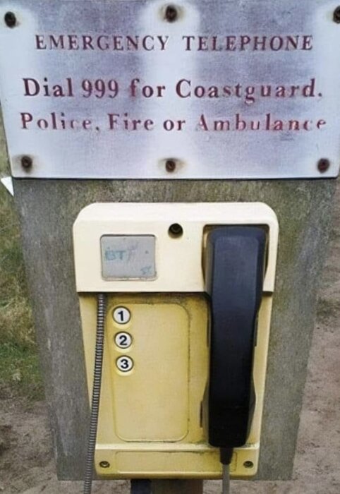 Nödtelefon med ett skylt ovanför, visar instruktioner att ringa 999 för kustbevakning, polis, brandkår eller ambulans. Telefonen har tre knappar märkta 1, 2, 3.