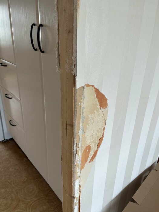 Uppsågad öppning i en vägg med skadad gips som behöver repareras. Väggen har delvis trasig tapet och avskalad färg. En vit garderob syns i bakgrunden.