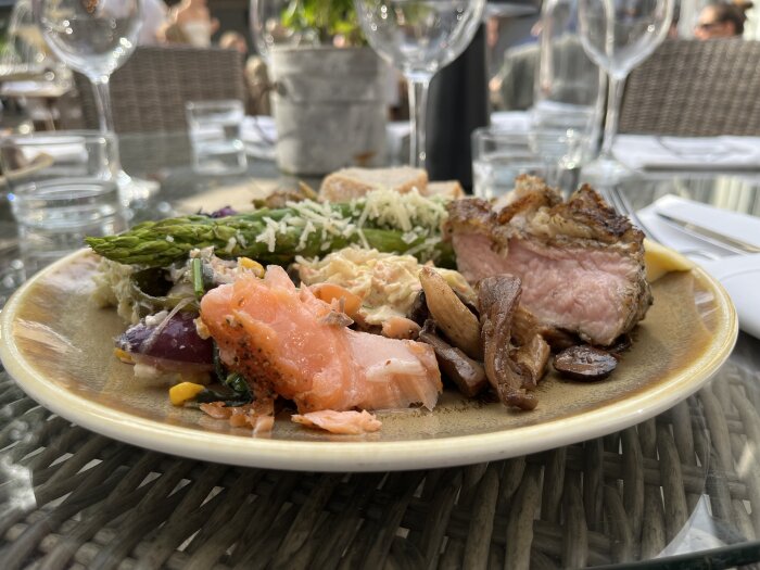 Tallrik med grillat kött, lax, sparris, svamp och pastasallad, serverad utomhus på en restaurang. Detaljer från en buffémiddag med glas i bakgrunden.