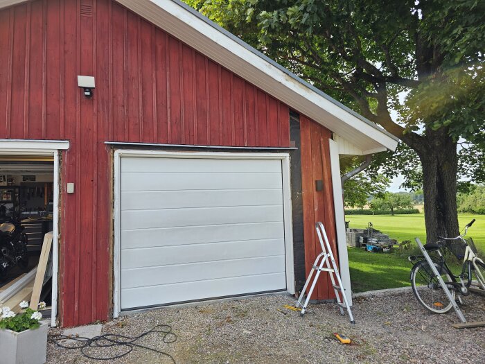 Rött garage med ny vit garageport, byte av panel pågår, stege längs väggen och delvis öppen dörr visar insidan med verktyg och motorcykel.