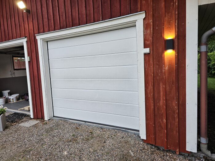 Rött garage med vitmålad garageport och ny bytt panel och foder. Grus framför garaget och ett påslaget utomhusljus på väggen.