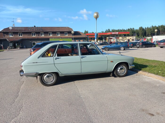 Ljusblå Renault 16 i utmärkt skick parkerad utanför ett hotell och en bensinstation med flera andra bilar runtom.