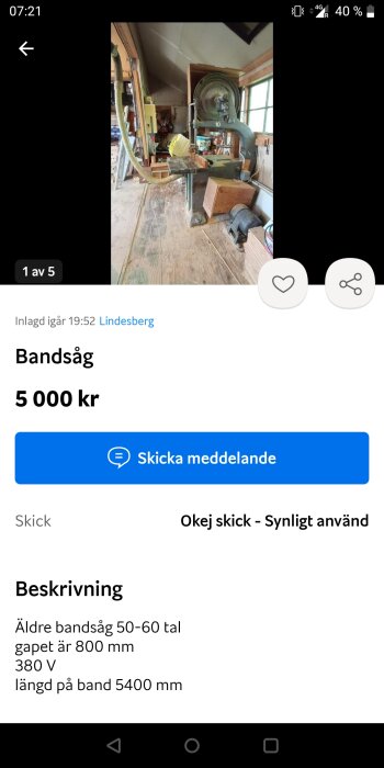 Äldre bandsåg i en verkstad, stående på ett trägolv med diverse verktyg och utrustning runtomkring.