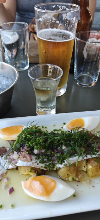 Bild av en förrätt med fisk, kokta ägg, hackad lök och örter på en tallrik, med öl och alkoholshotglas i bakgrunden.