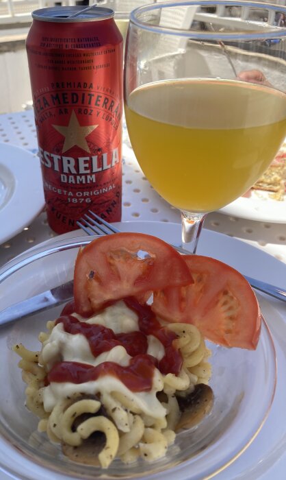 En burk Estrella Damm-öl, ett vinglas med öl, och en tallrik med pastasallad, sås och tomater på ett bord.