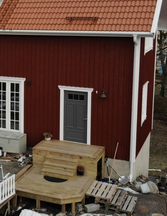 Röd trävilla med svart dörr och trappa av trä i två nivåer. Entrén saknar tak och staket som planeras att byggas av ägarna. Snözon 2.