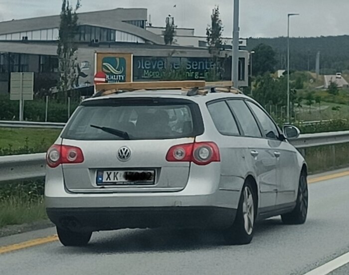 Silverfärgad Volkswagen kombi med träbitar på taket på motorväg i Norge, byggnad och skog i bakgrunden. Norsk registreringsskylt.