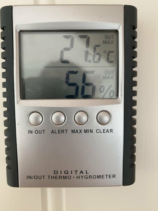 Digital hygrometer visar 27,6 grader Celsius och 56% luftfuktighet. Apparaten har knappar märkta "IN-OUT," "ALERT," "MAX-MIN" och "CLEAR" samt texten "DIGITAL IN/OUT THERMO·HYGROMETER.