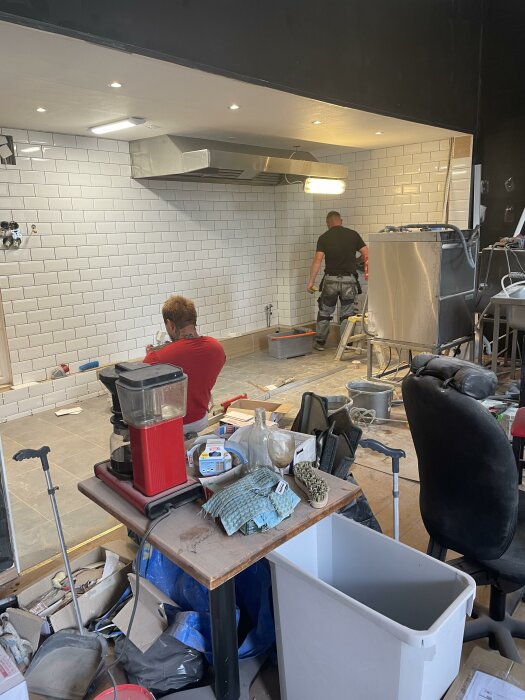 Två personer arbetar med att kakla en vit vägg i ett café under renovering; en sitter på golvet vid arbetsbordet fullt med verktyg, den andra står vid väggen.