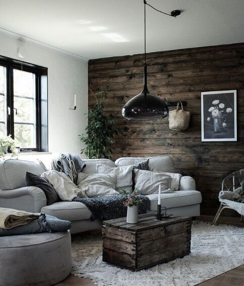 Rustikt vardagsrum med vit soffa, soffbord av mörkt trä och träpanel på väggen. Svart taklampa hänger ovanför bordet. Texturerade kuddar och filtar på soffan.