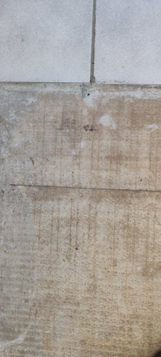 Klinkergolv möter en yta med spånskiva, där det saknas klinker och en dörrmatta legat. Ett parti av betong eller underlagsmaterial syns.