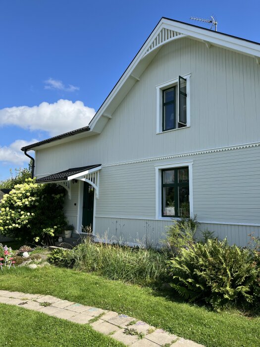 Exteriör av ett vitt hus med nytt skärmtak över dörren, omgärdat av en trädgård med buskar och gräs samt en blå himmel i bakgrunden.