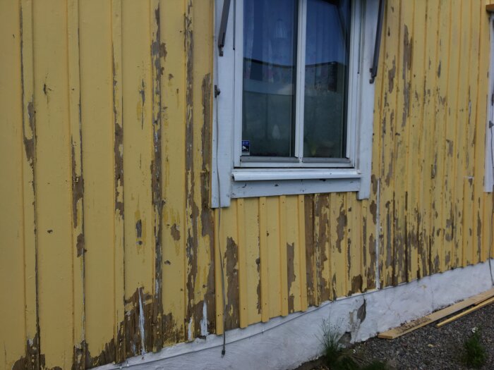 Gul träfasad med avskavd färg runt ett fönster; fogmassa applicerad vid vissa sömmar. Förberedelser inför tilläggsisolering och ny fasad.