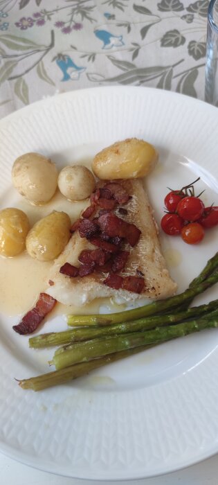 En tallrik med stekt torsk toppad med bacon, ångade potatisar, sparris och körsbärstomater på en vit duk med blommigt mönster.