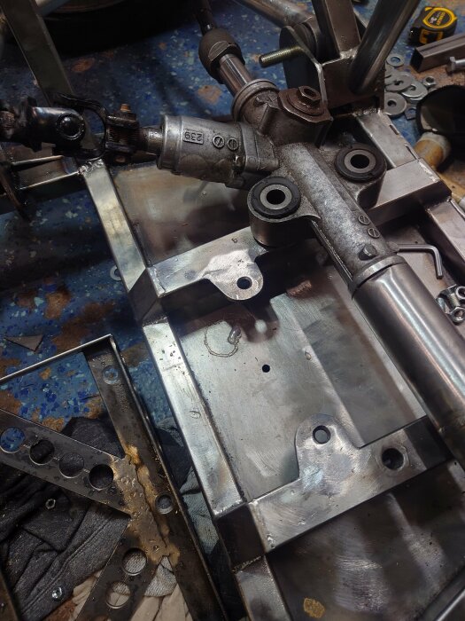 En svetsad metallkonstruktion med fästen för en styrkomponent ligger på ett verkstadsbord. Skruvar och metallbrackets är utspridda runt om.