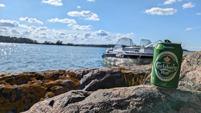 En Carlsberg Hof-burk står på en sten vid vattnet med en motorbåt i bakgrunden under en solig dag.