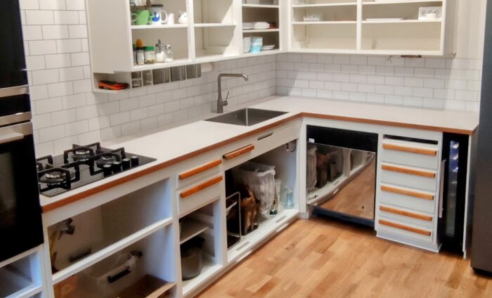Kök under renovering med öppna underskåp, inbyggd diskmaskin utan front längst ner till höger och färdigmonterad bänkskiva, vit kakel och trägolv.