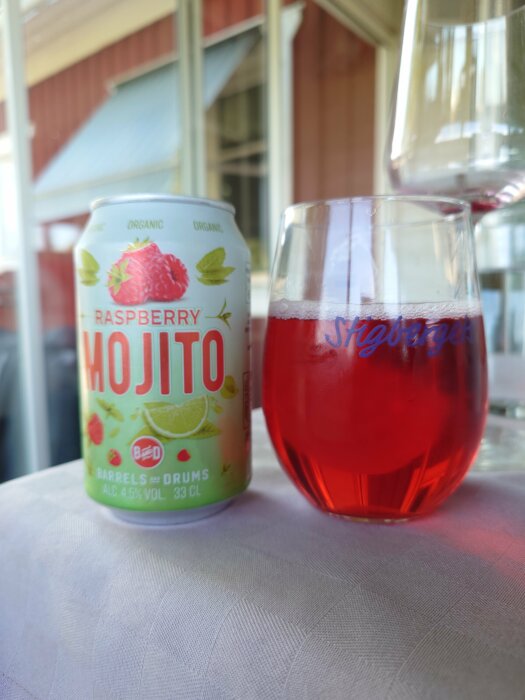 En burk med Raspberry Mojito och ett fyllt glas på ett bord, med ett hus i bakgrunden.