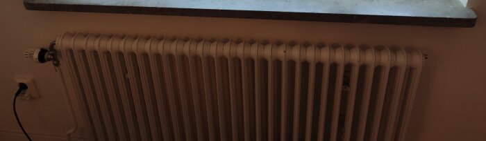 Radiator under ett fönster i ett rum, med en termostat på vänster sida kopplad till ett rör. En eluttag med en kontakt ligger till vänster om radiatorn.