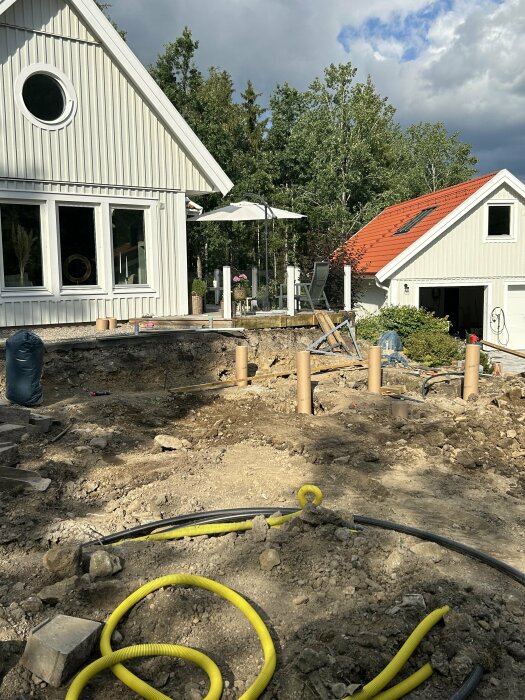 Byggarbetsplats med grävd mark för plintar, flera plintar synliga i marken, hus i bakgrunden och träaltan med parasoll. Gul elkabel på marken.