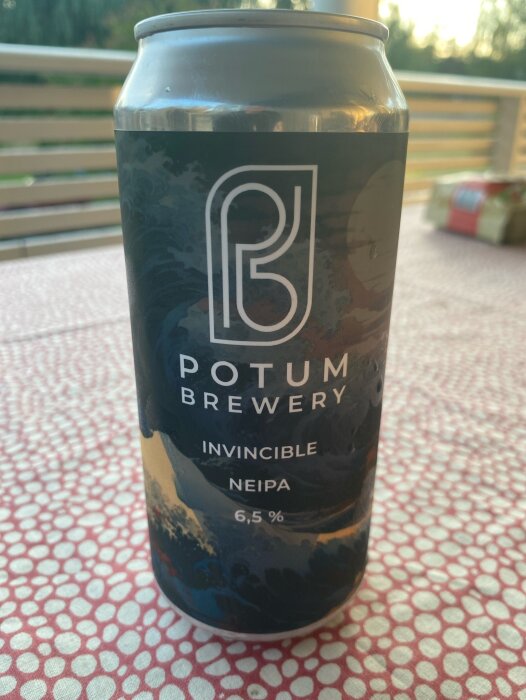 En ölburk från Potum Brewery med etiketten "Invincible Neipa 6,5%" på ett bord med mönstrad duk.