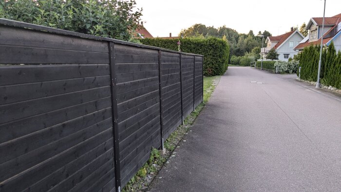 Svartmålat träplank som sträcker sig längs en trottoar i ett villaområde. Planket har funnits i 14 år och är behandlat med linolja.