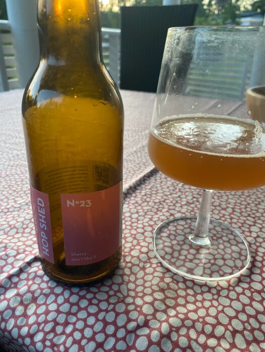 En flaska Hop Shed No 23 öl står på ett bord bredvid ett halvtomt glas med grapefruit IPA. Bakgrunden visar en uteplats.