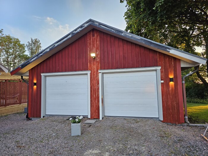 Rött garage med två vita garageportar, grusuppfart och lampa tänd på väggen. Röd träpanel där några brädor är utbytta. Träd och gräsmatta i bakgrunden.